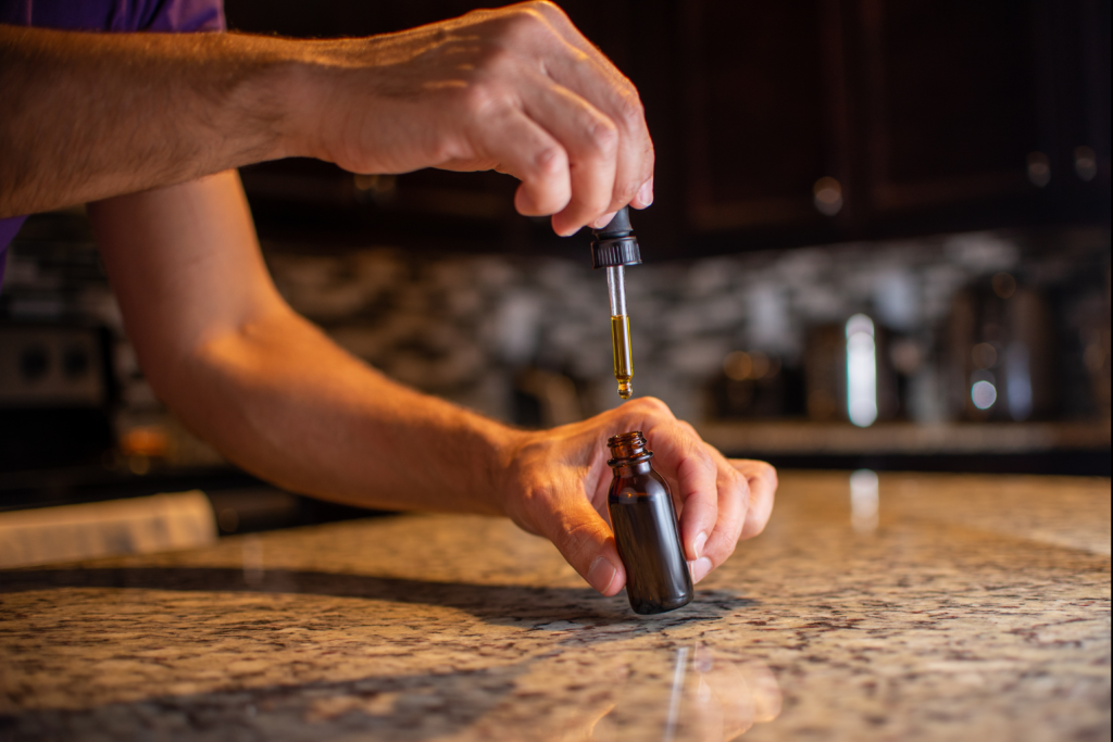 Close-up photo of a person using a dropper to dispense amber tincture oil into a small brown glass bottle on a kitchen countertop, with a softly blurred background.