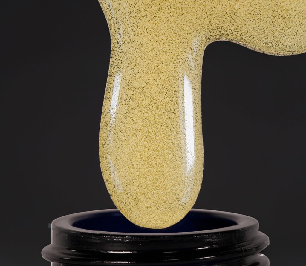 Macro close-up of golden, terpene-rich hash rosin stretching downward in a thick glossy drip into the opening of a black jar. The concentrate has a granular, crystalline texture with bright highlights against a dark background.