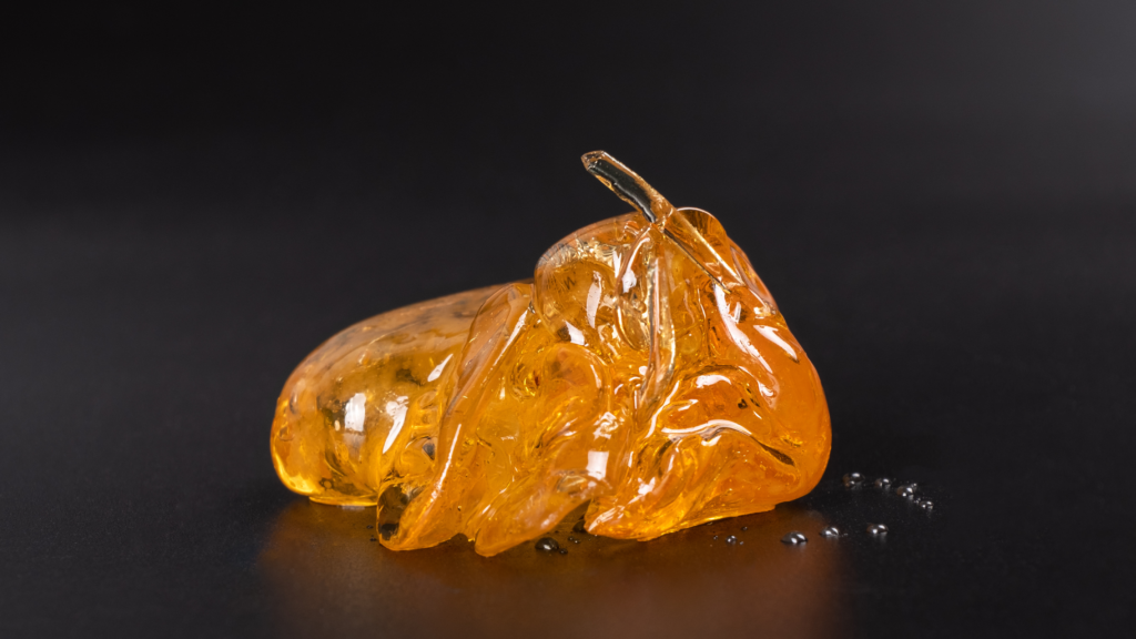 Macro image of translucent amber cannabis concentrate with a glossy, glass-like texture resting on a dark surface. Small droplets and reflections surround the golden extract against a black background.