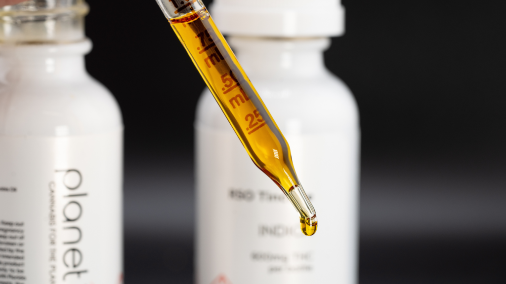 Close-up of a glass dropper filled with golden cannabis oil, with a small droplet forming at the tip. Blurred white Planet 13 tincture bottles sit in the background against a dark backdrop.
