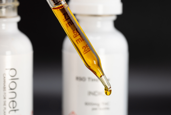 Close-up of a glass dropper filled with golden cannabis oil, with a small droplet forming at the tip. Blurred white Planet 13 tincture bottles sit in the background against a dark backdrop.