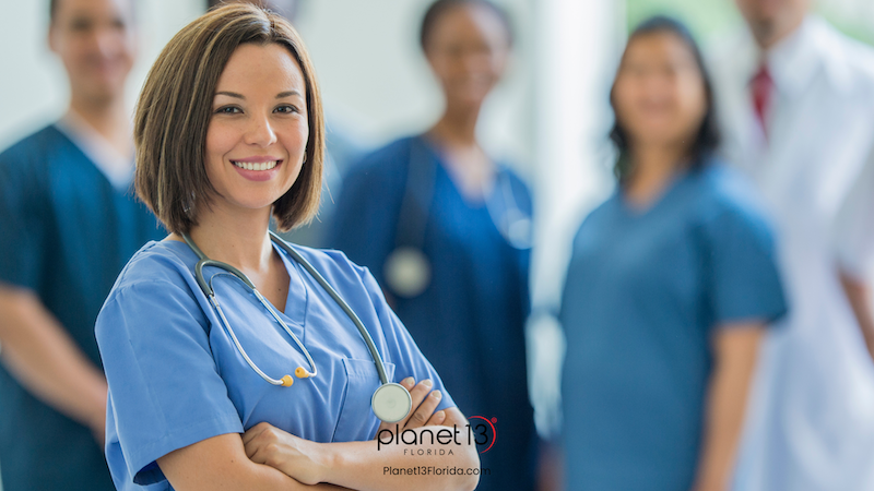 Smiling female doctor standing in a healthcare setting with colleagues behind her.