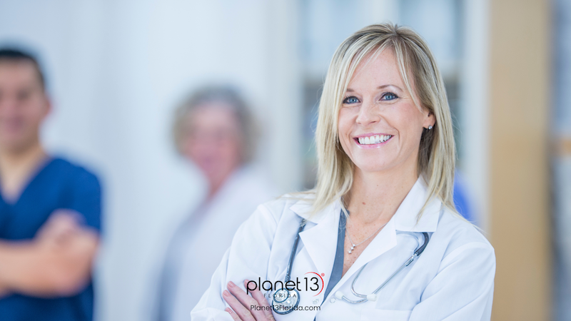 Smiling female doctor wearing a white coat with a blurred colleague behind her.