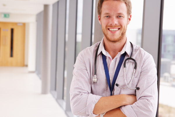Smiling male doctor wearing a white coat and stethoscope in a clinic hallway.