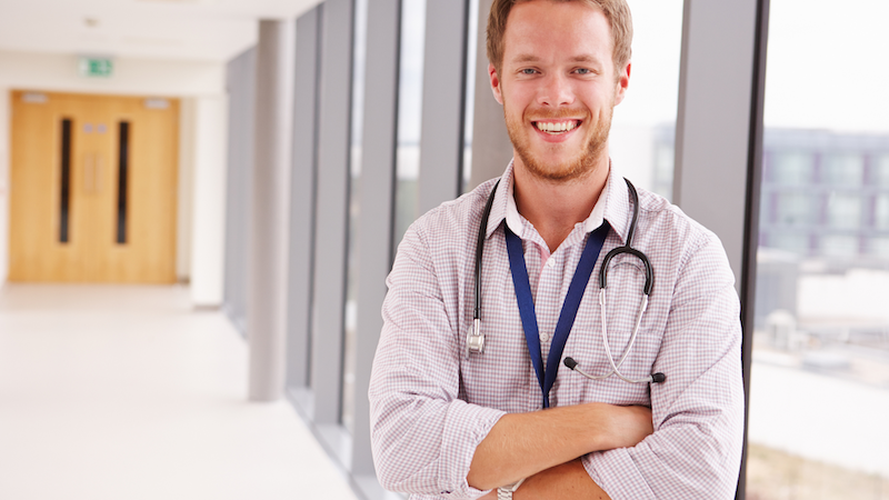 Smiling male doctor wearing a white coat and stethoscope in a clinic hallway.